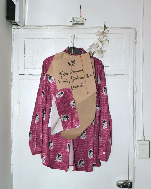 A fuschia shirt adorned with face graphics hangs on a white door, accompanied by a hand-written cardboard sign and white orchids.