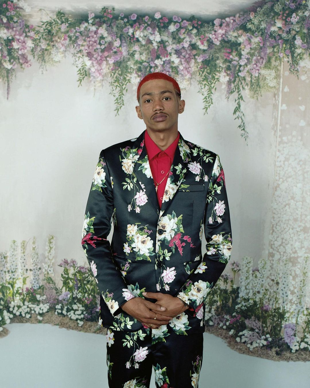 Man in a floral suit stands formally in front of a floral backdrop. He is wearing a red shirt and gold chain. 