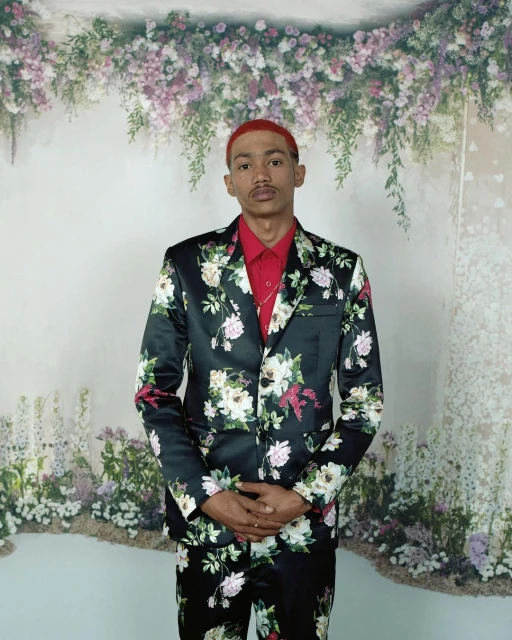 Man in a floral suit stands formally in front of a floral backdrop. He is wearing a red shirt and gold chain. 