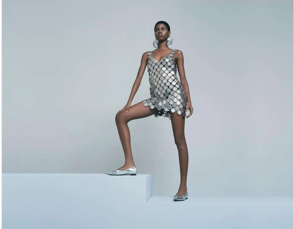Model wearing a metallic disc-embellished mini dress and silver flats posing on a block in a studio