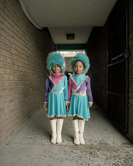 Two young black girls in matching colorful costumes, including turquoise dresses and large fur hats, stand in a hallway, posing for the camera.