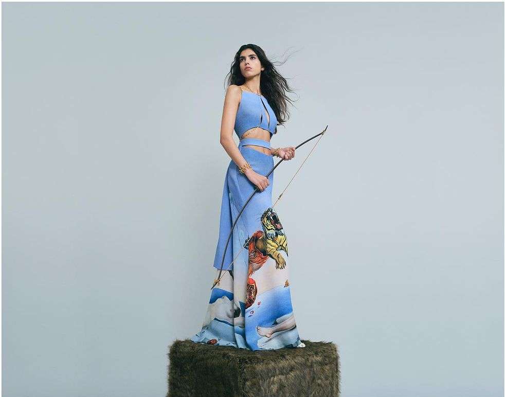 A woman poses wearing a blue two-piece dress with a bow and arrow. The dress is printed with art of a tiger and human figures.