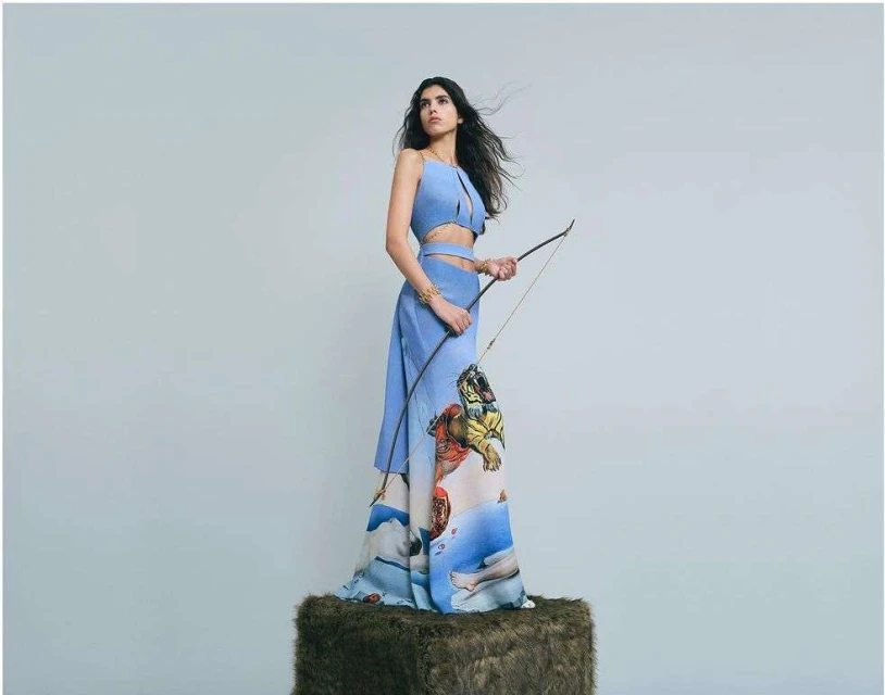 A woman poses wearing a blue two-piece dress with a bow and arrow. The dress is printed with art of a tiger and human figures.