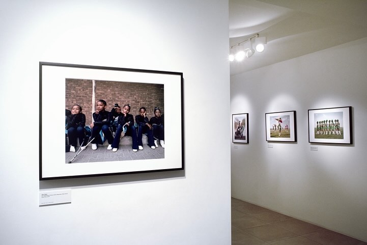 Art gallery wall displaying a photograph of a group of schoolgirls sitting on a ledge in matching uniforms against a brick wall backdrop.