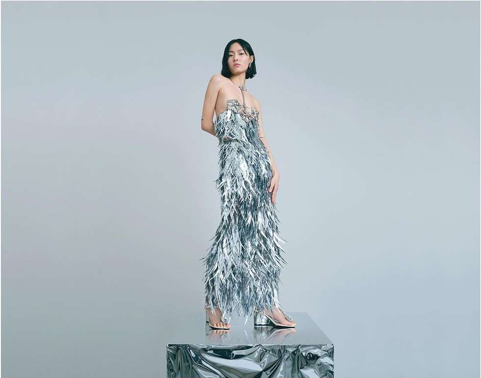 Young woman posing in silver feathered dress on a metallic platform in a photo studio setting. Fashion editorial shot.