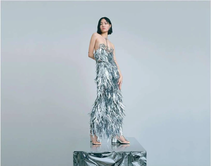 Young woman posing in silver feathered dress on a metallic platform in a photo studio setting. Fashion editorial shot.