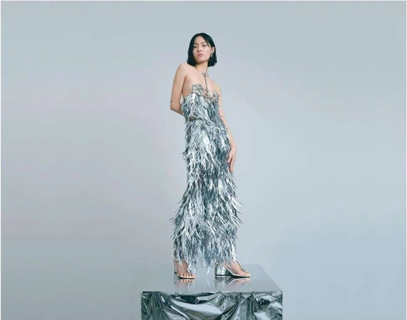 Young woman posing in silver feathered dress on a metallic platform in a photo studio setting. Fashion editorial shot.