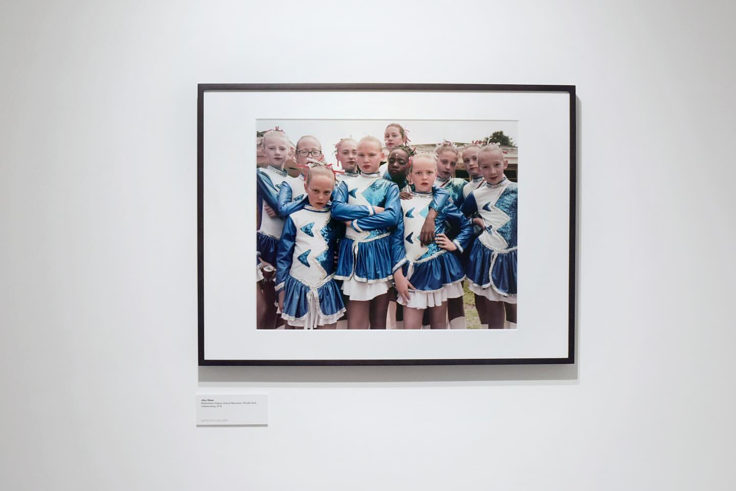 A framed photograph displays a group of young girls in matching blue and white costumes posing together outdoors with neutral expressions.