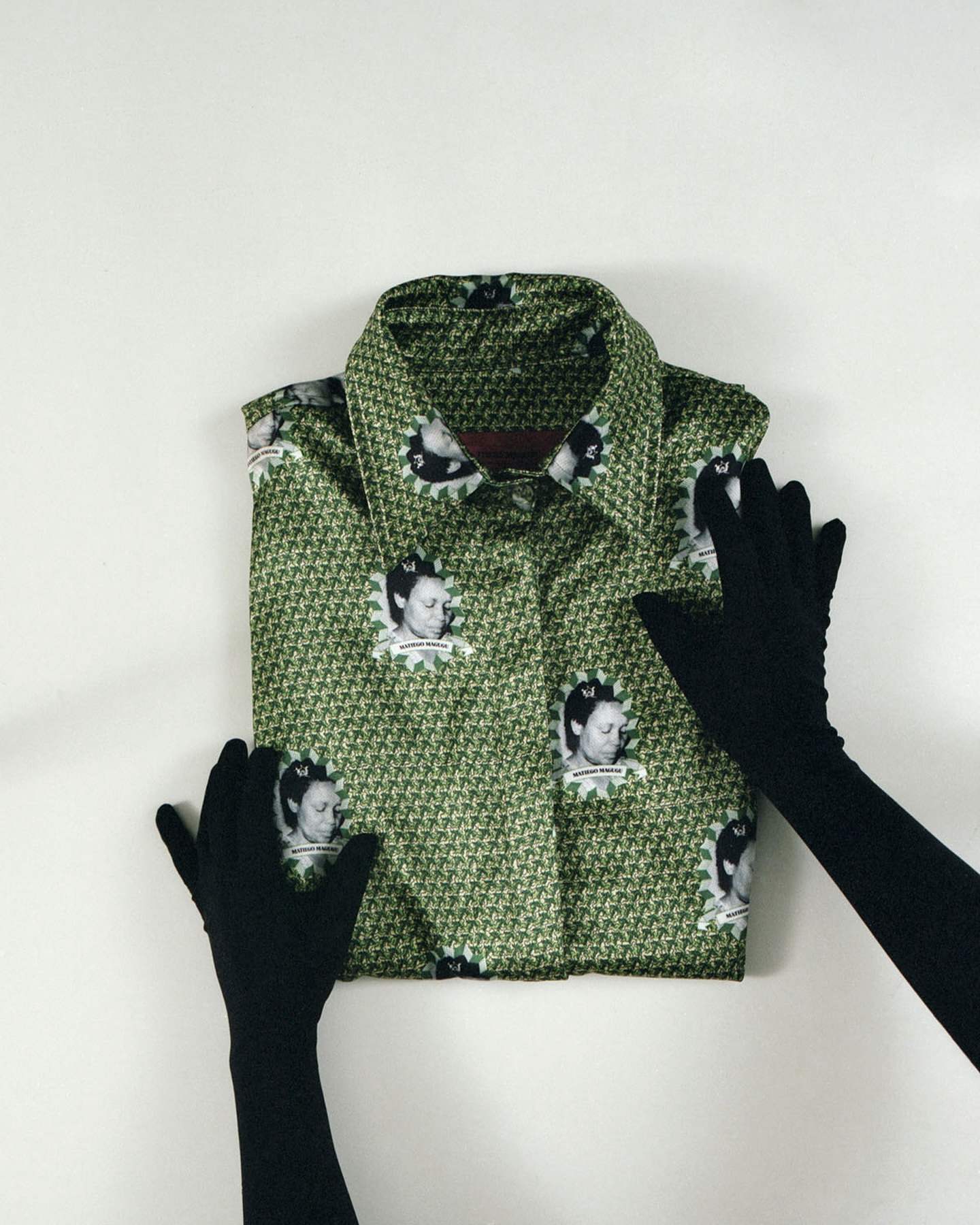 Green collared shirt with portraits pattern being held by black gloved hands on a white surface in a studio-like setting.