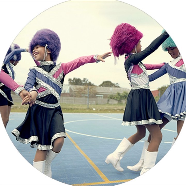 Several young girls wearing matching performance outfits, including knee-high white boots and large colorful fuzzy hats, on an outdoor court.