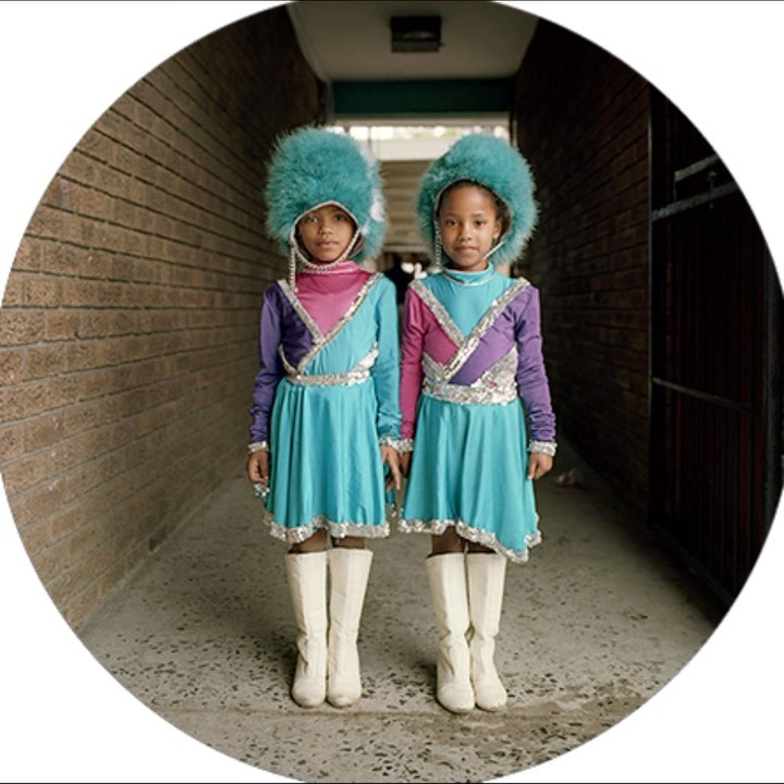 Two young girls in matching turquoise dresses with ornate silver trim and white boots standing in a hallway with brick walls.