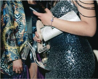 Two people at an event, one wearing a sparkly sequin dress holding clear heeled sandals and a silver clutch.