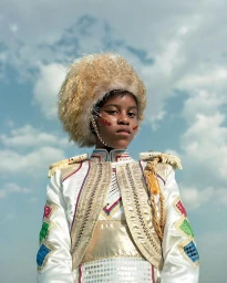 Young Black person wearing ornate white and gold ceremonial costume with a large fluffy blonde hat against blue cloudy sky