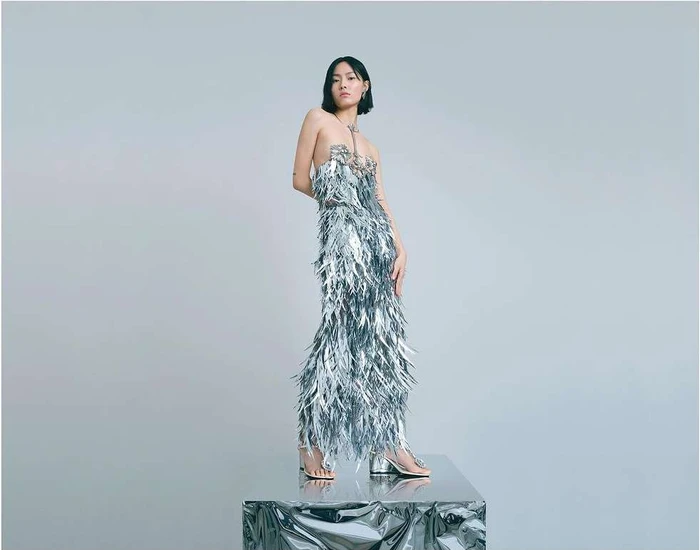 East Asian woman wearing a metallic fringed halter dress standing on a reflective surface against a plain grey background