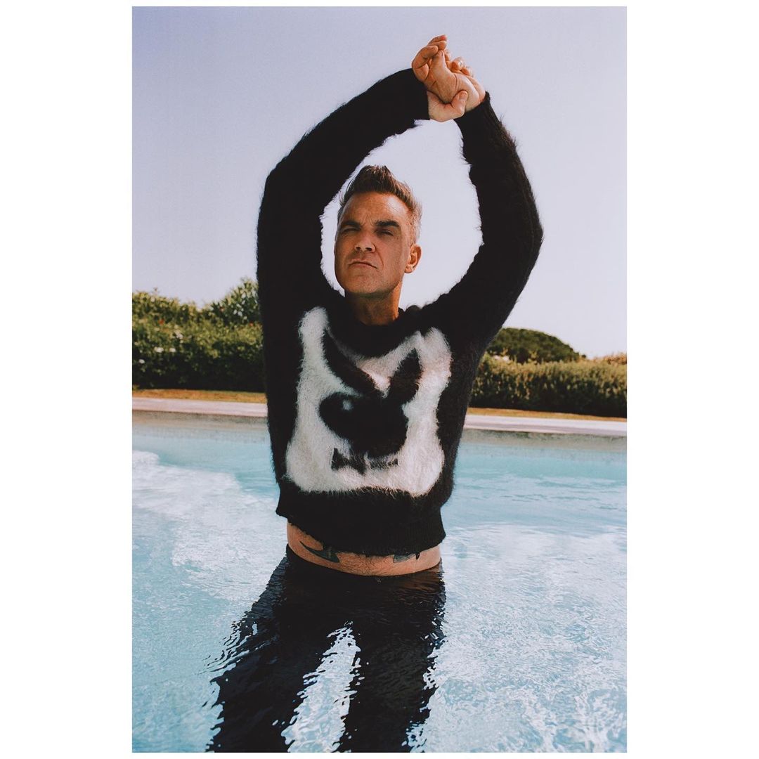 Adult male standing in a pool, wearing a black sweater with white rabbit logo, arms raised, and outdoor setting. Fashion.