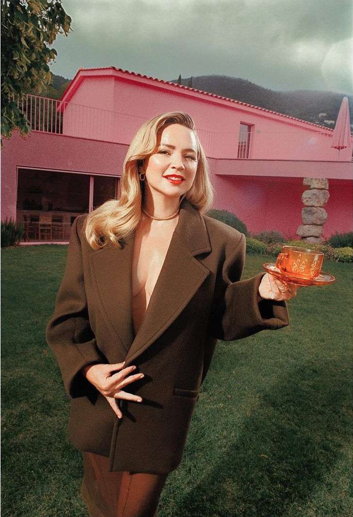 Woman poses outdoors in an olive blazer and skirt, holding a tea cup and saucer, with a pink house and green trees in background.