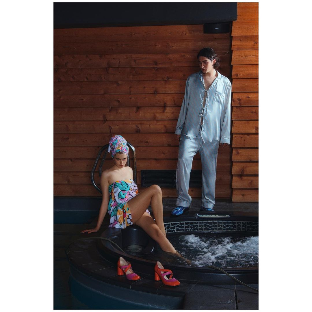 Woman in a patterned towel and pink shoes sits near a jacuzzi while a man in pajamas stands in the background in a wood-lined room.
