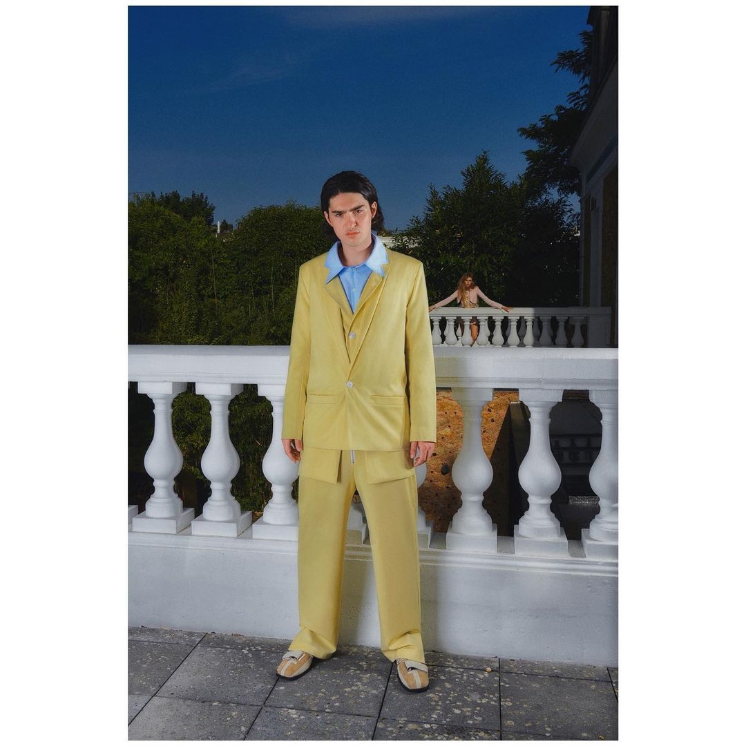 A man in a yellow suit stands near a white stone balustrade with another person behind it, under a clear blue sky and green trees.