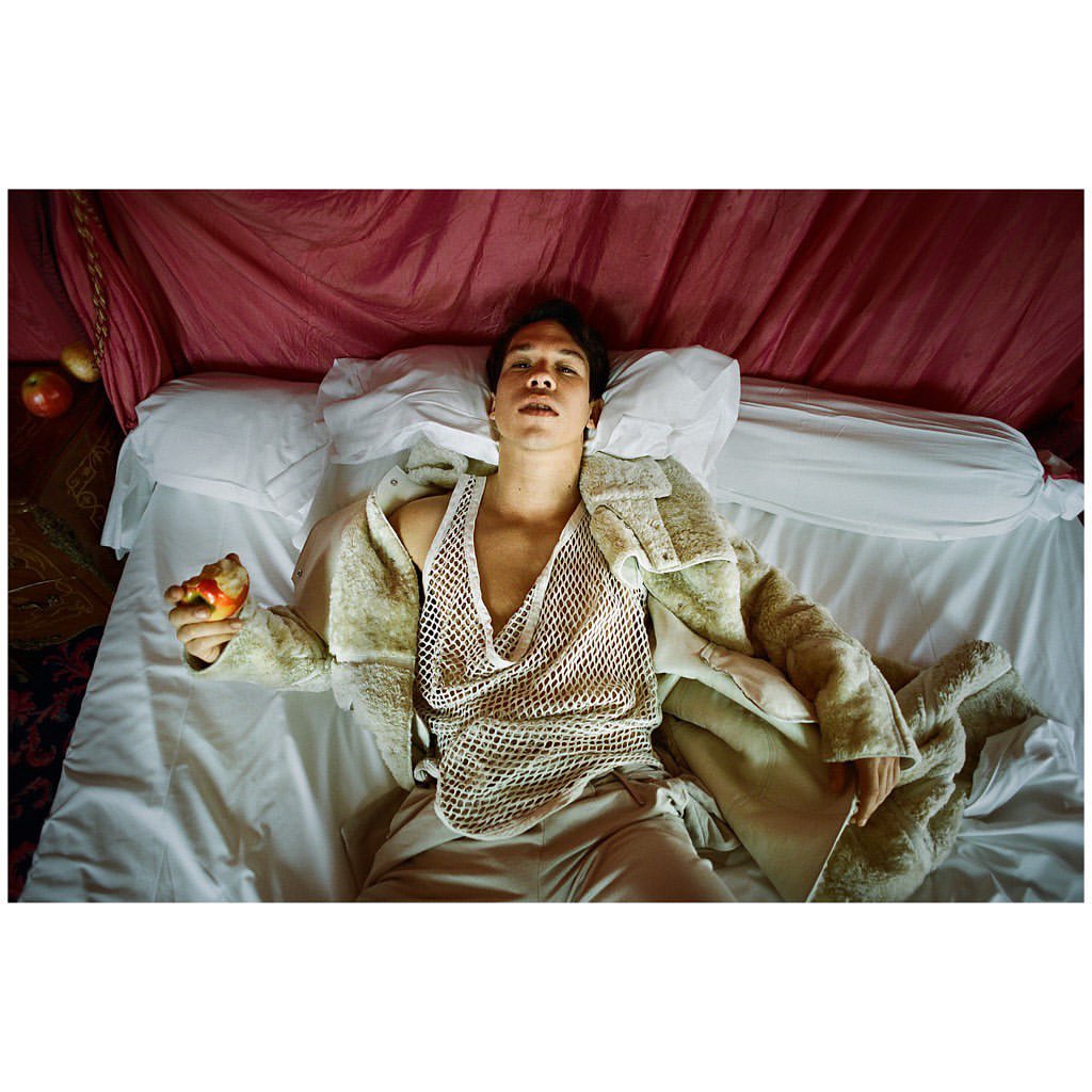 Young Asian man lies on a bed with white linens, wearing a fishnet shirt and fur coat, holding an apple, with a red canopy above.