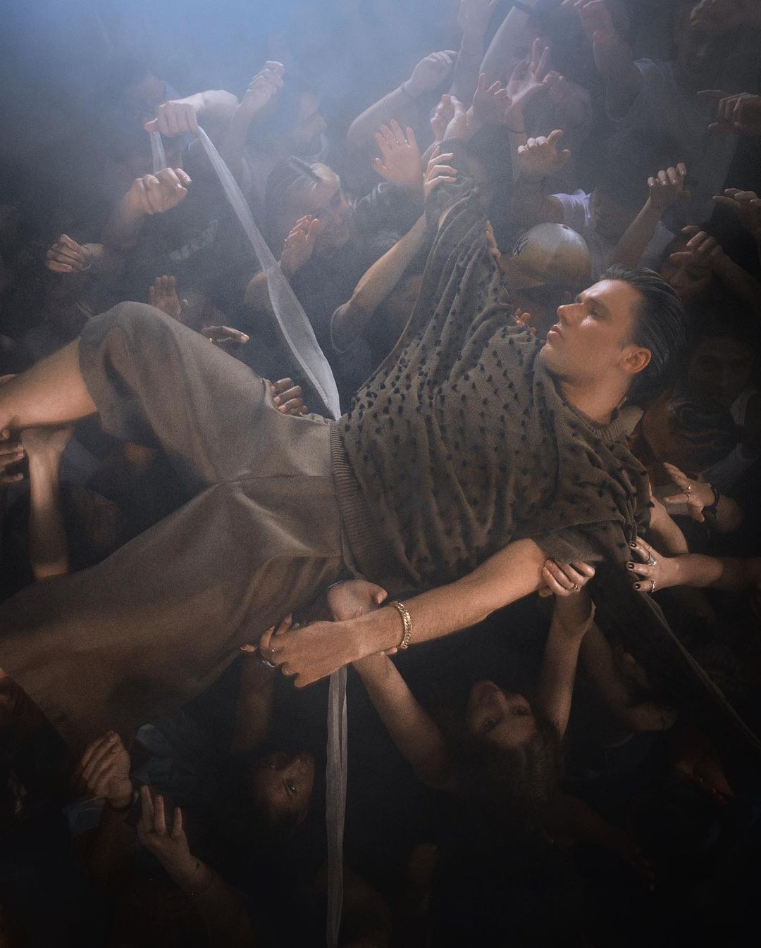 Man lying down surrounded by reaching hands in a dark, moody scene. The image evokes a sense of drama and vulnerability.