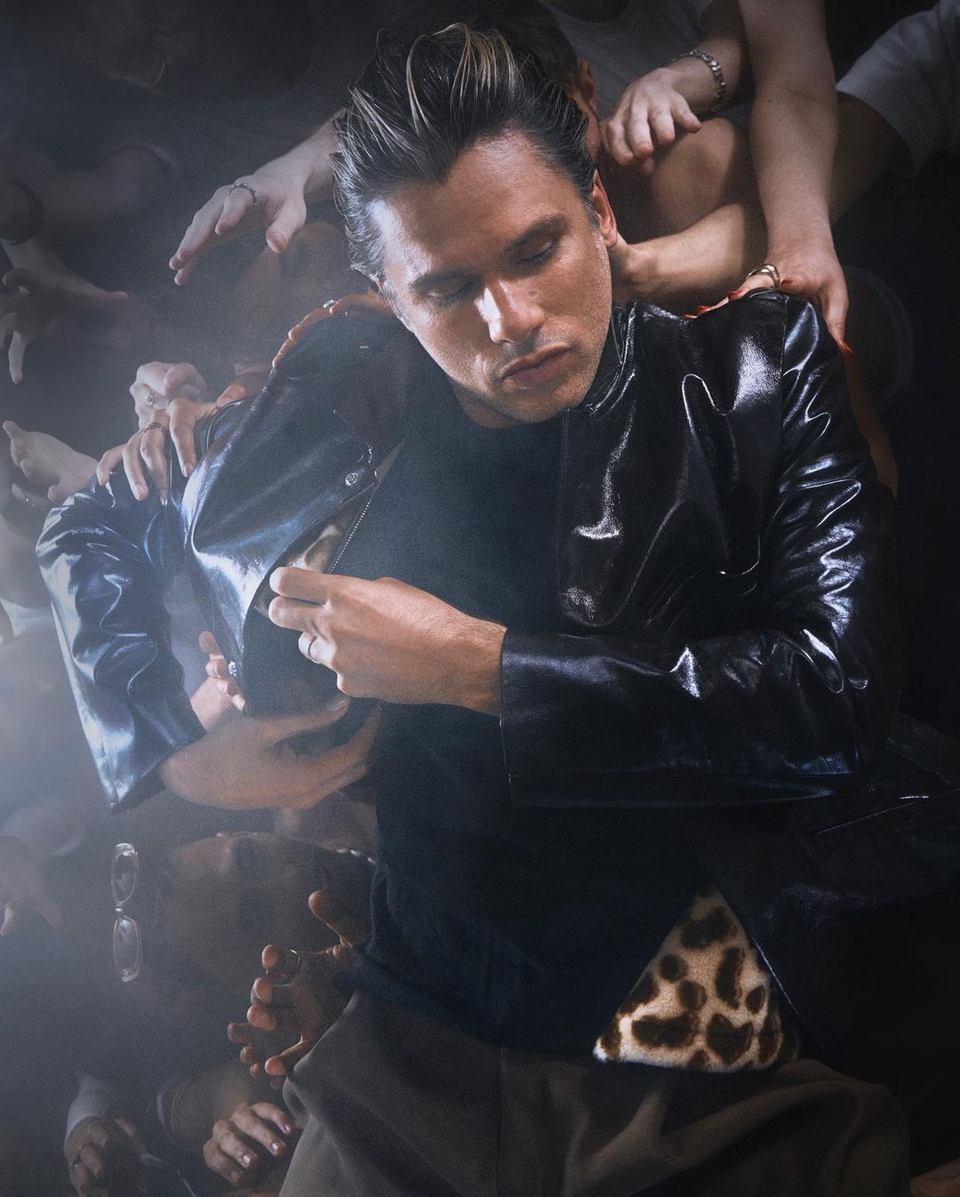 Portrait of a man in a black jacket, surrounded by a crowd of hands, with a dark background and soft, diffused lighting.