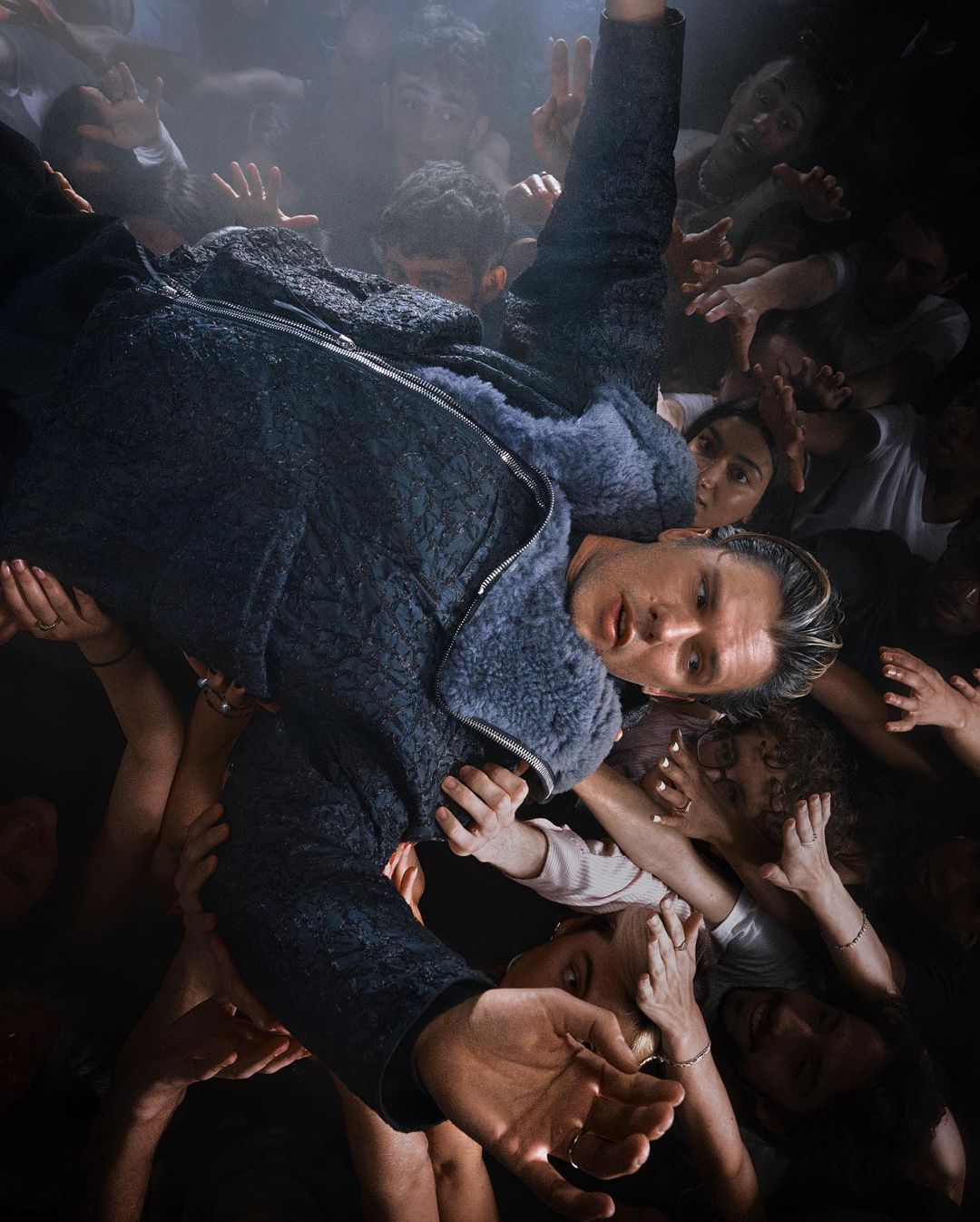 Person crowd surfing with many hands reaching up. Adult male with blond hair and blue jacket in a dark indoor environment.