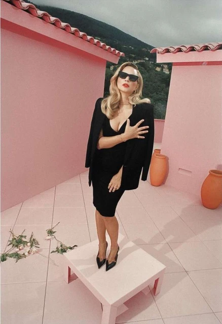 Elegant woman in sunglasses stands on a pink platform in a black dress with a jacket, roses and terracotta jugs, set against a mountain backdrop.