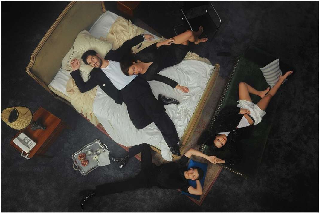 Overhead view of four adults in formal wear lying on beds and floor in a dark room with neutral colors and subdued lighting.