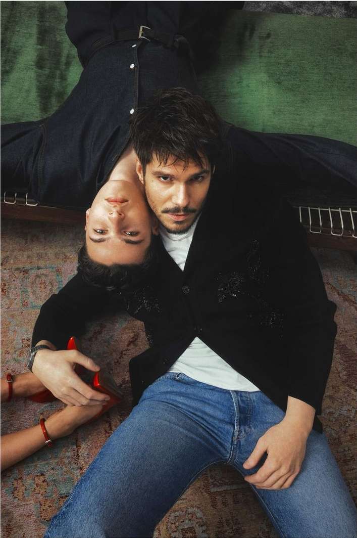 Two adults lying on a patterned rug indoors. One adult wears blue jeans and a black cardigan. The other adult wears a denim jumpsuit.