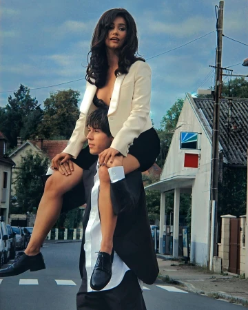 A woman riding on a man's shoulders on a street, wearing a white blazer and dark top, photographed by Alice Moitié.