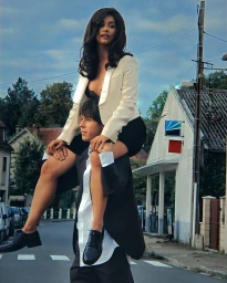A woman riding on a man's shoulders on a street, wearing a white blazer and dark top, photographed by Alice Moitié.