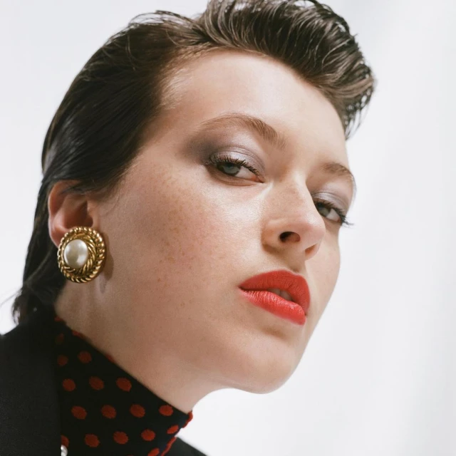 Close-up studio shot of a young, fair-skinned adult female with red lipstick and gold pearl earrings, against a white background.