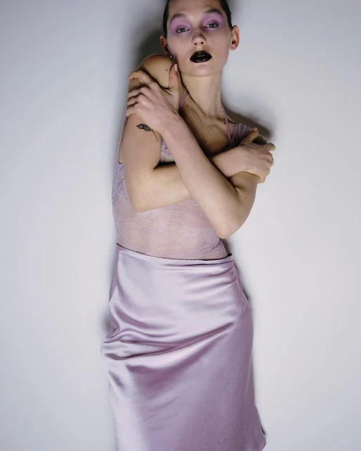 Portrait of a slender adult female wearing a pale pink slip dress and dark lipstick with her arms crossed in an indoor studio setting.