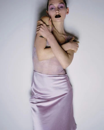 Portrait of a slender adult female wearing a pale pink slip dress and dark lipstick with her arms crossed in an indoor studio setting.
