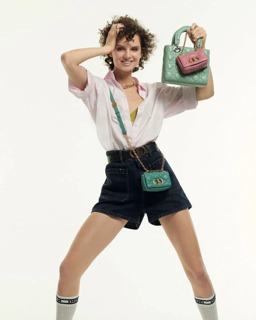 Fashionable woman in shorts holds up a green quilted handbag and pink mini handbag in front of a plain white background.
