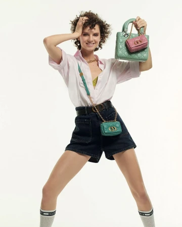 Fashionable woman in shorts holds up a green quilted handbag and pink mini handbag in front of a plain white background.