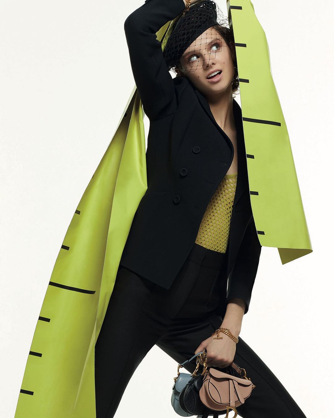Fashion editorial shot of a young woman wearing a black blazer, trousers and net hat, holding two handbags against a light backdrop.