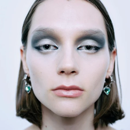 Close-up of a woman's face with smokey gray eye makeup and glossy lips, wearing dangling heart-shaped earrings, against a white background.