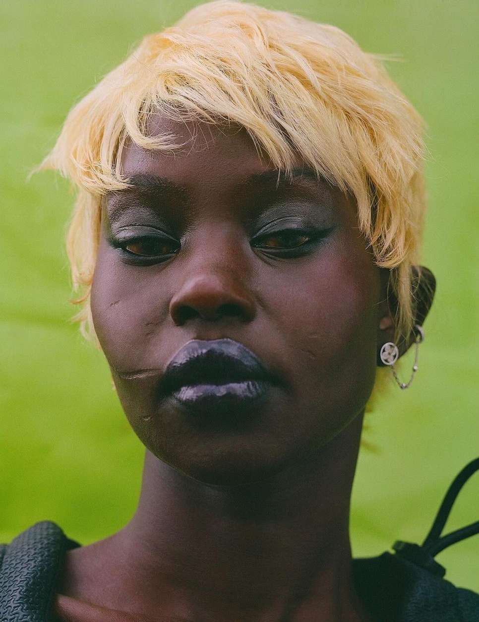 Close-up portrait of a dark-skinned woman with short blonde hair and a neutral expression against a lime-green background.