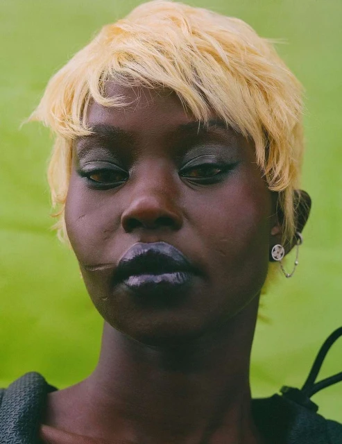 Close-up portrait of a dark-skinned woman with short blonde hair and a neutral expression against a lime-green background.