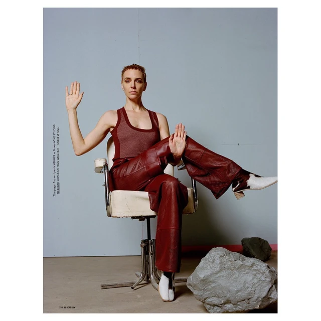 Adult woman poses in brown leather pants and top, sitting in a chair in a photo studio with rocks in the foreground.