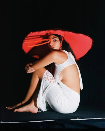 Model wearing a wide red sun hat and white cut-out dress seated against a dark black background, photographed in Paris.