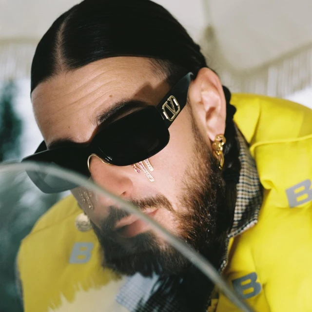 Close-up of a stylish adult male with a beard, sunglasses, and yellow puffer jacket, adorned with jewelry, in an indoor setting.