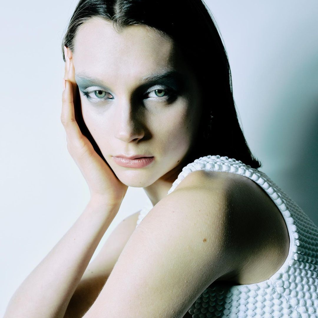 A portrait of an adult female model with creative makeup and a white textured dress is set against a soft, neutral background.