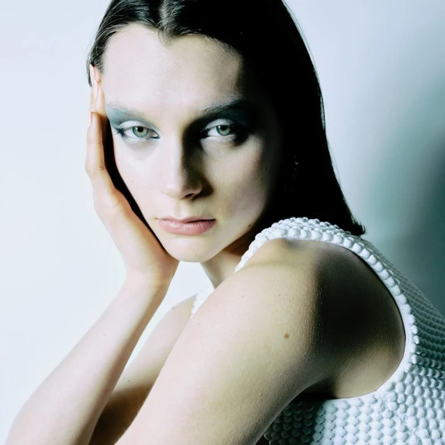 A portrait of an adult female model with creative makeup and a white textured dress is set against a soft, neutral background.