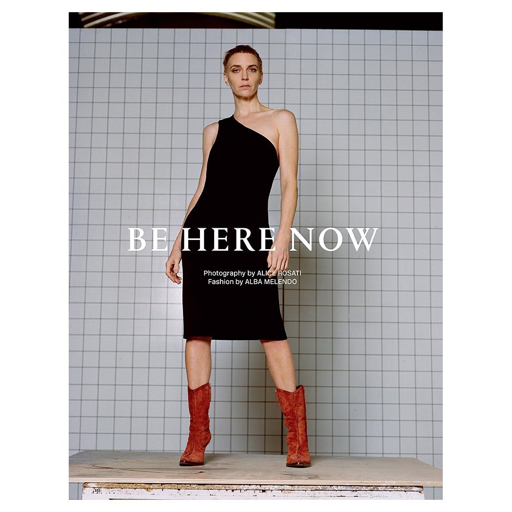 Fashion editorial shot of a model in a black dress and red boots, posing against a tiled wall. Studio lighting emphasizes the outfit and design.