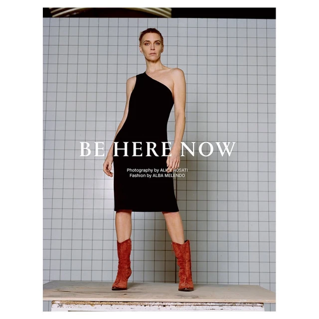 Fashion editorial shot of a model in a black dress and red boots, posing against a tiled wall. Studio lighting emphasizes the outfit and design.