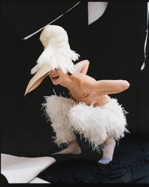 An adult poses in an artful, theatrical headpiece and feathery costume, with exaggerated expressions, against a black backdrop.