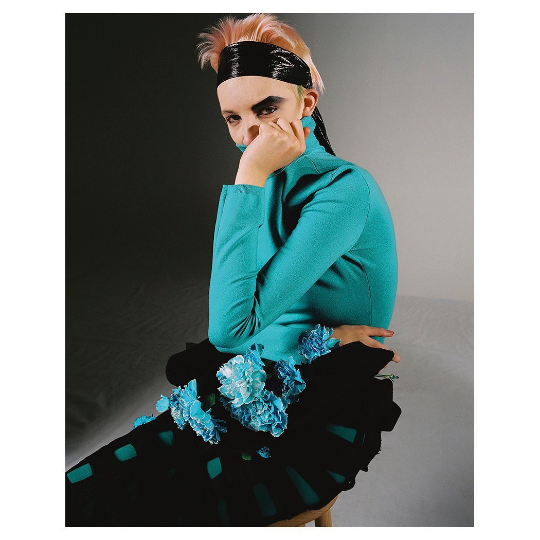 Portrait of a stylish adult female wearing a turquoise turtleneck and black skirt with blue flowers, headband, and makeup against a grey background.