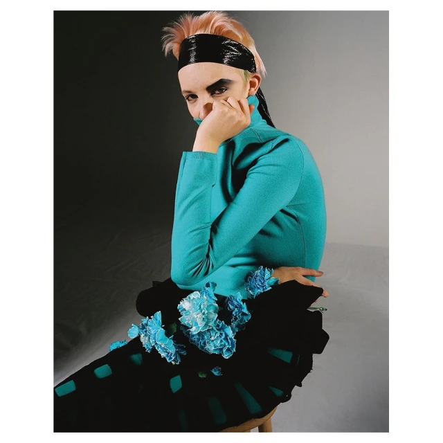 Portrait of a stylish adult female wearing a turquoise turtleneck and black skirt with blue flowers, headband, and makeup against a grey background.
